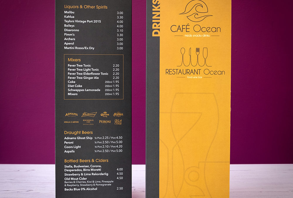 Photograph of a tall A4 folding Cafe Ocean drinks menu standing on white wooden floorboards with a purple wall in the background