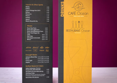 Photograph of a tall A4 folding Cafe Ocean drinks menu standing on white wooden floorboards with a purple wall in the background