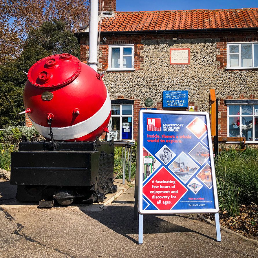 New A-board and promotional posters on the pavement outside the flint faced Lowestot Maritime Museum. The A-board is positioned next to bright red and white wartime sea mine.